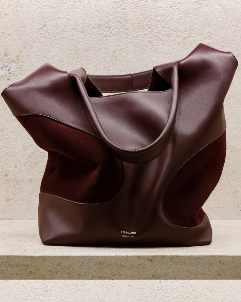 A Ferragamo handbag in a deep burgundy color, made of leather with curved panel designs. The bag is displayed on a light stone surface, highlighting its luxurious texture and craftsmanship.